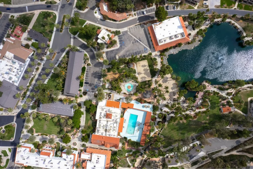Aerial view of a resort area with various buildings, palm trees, a swimming pool, landscaped pathways, and a pond. The image shows a well-organized layout with a mix of structures and natural elements, including greenery and water features.