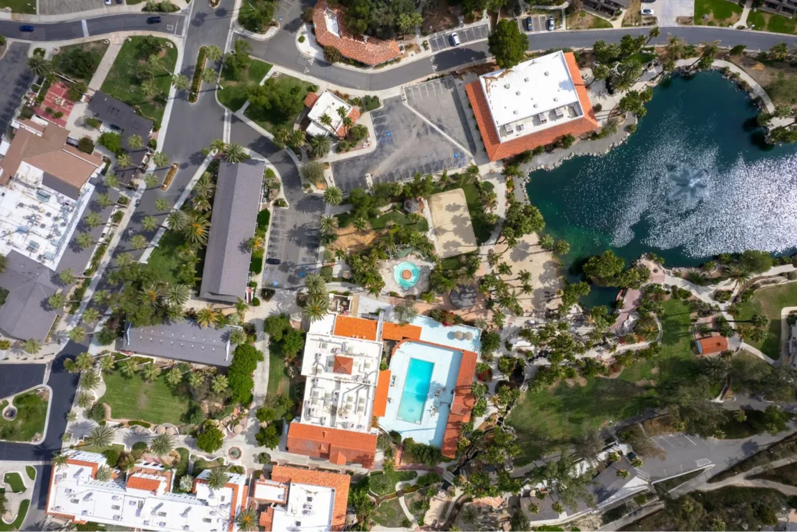 Aerial view of a resort area with various buildings, palm trees, a swimming pool, landscaped pathways, and a pond. The image shows a well-organized layout with a mix of structures and natural elements, including greenery and water features.